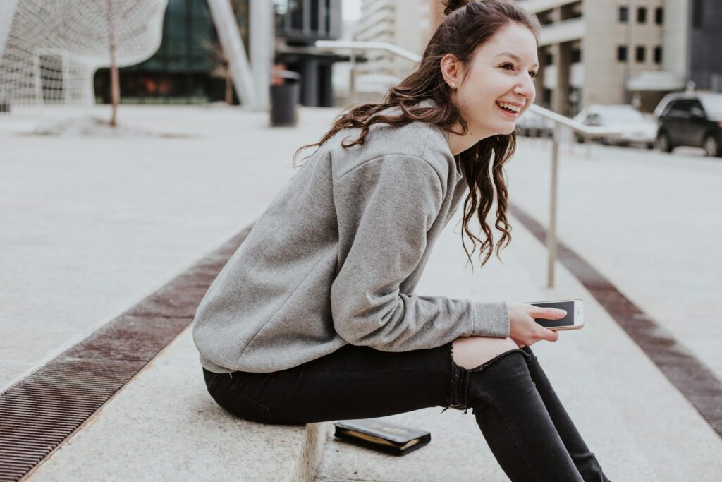 woman sits on sidewalk holding smartphone while smiling